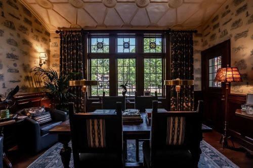 Cozy room with a wooden table, chairs, and large windows overlooking greenery, adorned with patterned wallpaper and lamps.