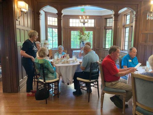 A group of seniors enjoying a meal together in a warmly lit dining room with wooden paneling and large windows.