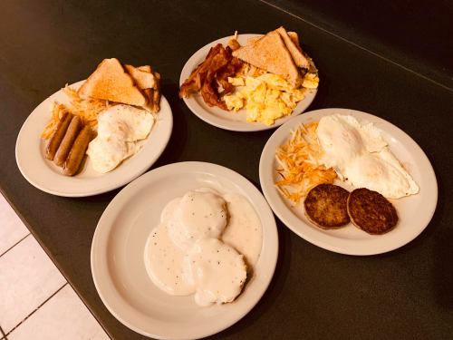 Four plates of breakfast: scrambled eggs, bacon, toast, sausage, hash browns, and biscuits with gravy.