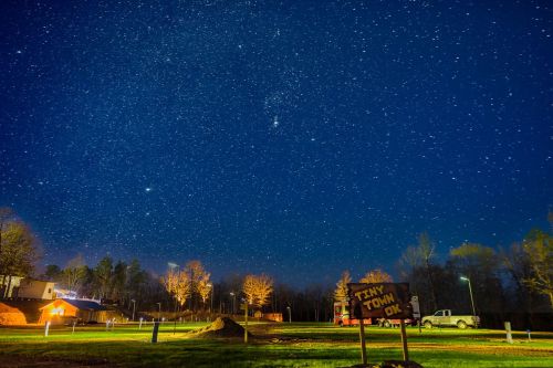 A starry night sky over a rural area, with trees, a sign reading "Tiny Town," and distant buildings illuminated.