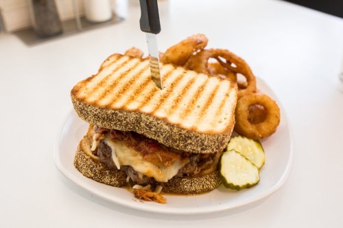 A hearty sandwich with grilled bread, melted cheese, and toppings, served with onion rings and pickles on a white plate.