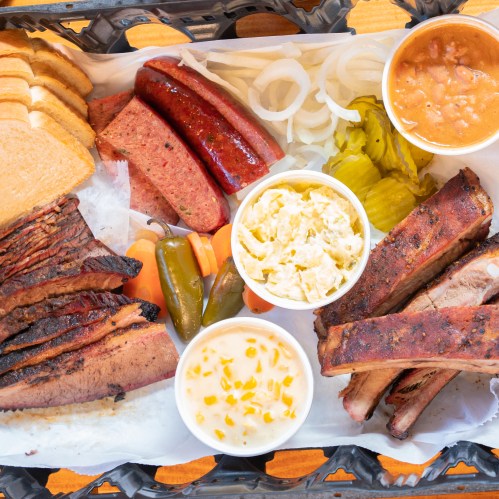 A platter of assorted meats including brisket, sausage, and ribs, served with sides of pickles, onions, and creamy dishes.