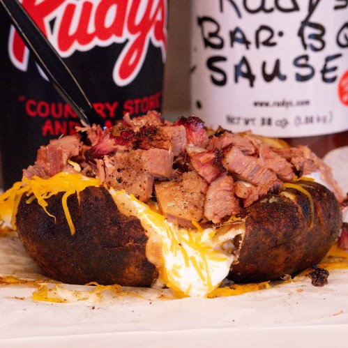 A close-up of a loaded baked potato topped with chopped barbecue meat and melted cheese, served in a paper tray.
