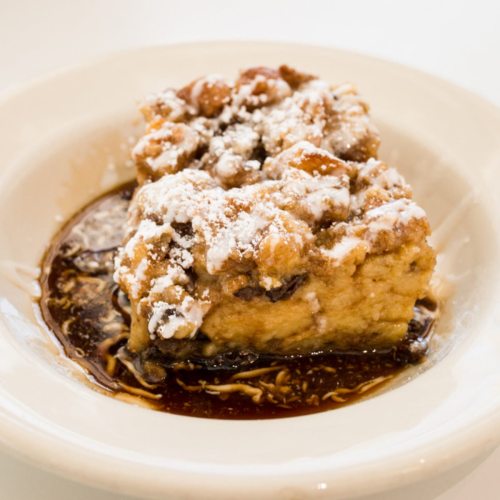 A slice of dessert topped with powdered sugar, served in a shallow dish with a dark syrup underneath.