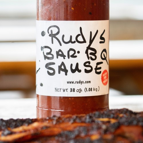 A bottle of Rudy's Bar-B-Q Sauce, 38 oz, with a white label and bold black text on a wooden surface.