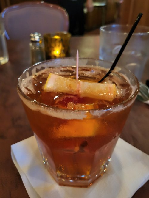 A close-up of a cocktail in a glass, garnished with a slice of fruit and a cherry, on a napkin at a restaurant table.