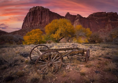 An old wooden wagon sits in a desert landscape with vibrant autumn trees and a dramatic sunset sky.
