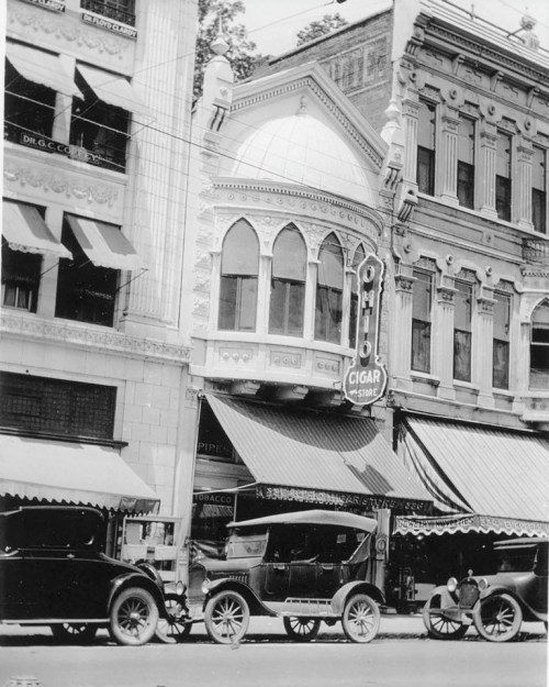 Historic street scene featuring vintage cars parked outside a cigar store with ornate architecture.