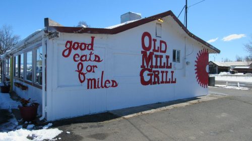 A white building with red lettering reading "Old Mill Grill" and "good eats for miles," surrounded by snow.