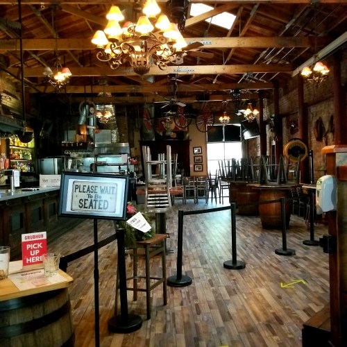 Interior of a rustic restaurant with wooden floors, chandeliers, and empty tables, featuring a "Please Wait to Be Seated" sign.