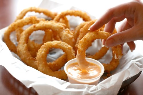 A hand dips a crispy onion ring into a small cup of sauce, surrounded by more onion rings in a basket.
