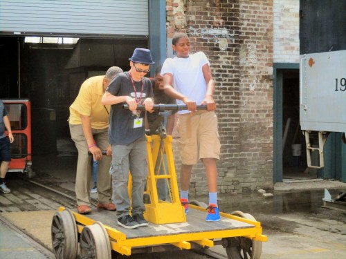 Two boys operate a yellow handcar on tracks, while an adult assists them in the background at a historic site.