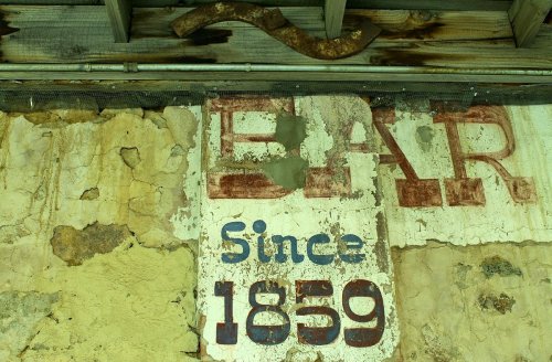Faded sign on a weathered wall reads "BAR" and "Since 1859," with peeling paint and rustic textures.