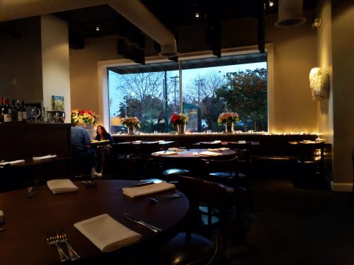 Cozy restaurant interior with tables set for dining, large windows, and floral decorations, illuminated by soft lighting.