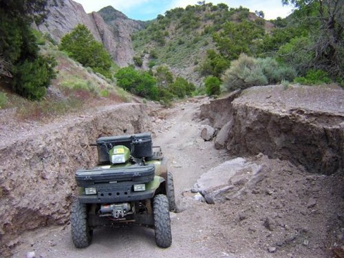 An ATV parked on a rugged, eroded dirt path surrounded by rocky terrain and greenery.