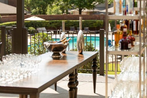 A table with champagne and glasses overlooks a pool area, surrounded by greenery and lounge chairs.