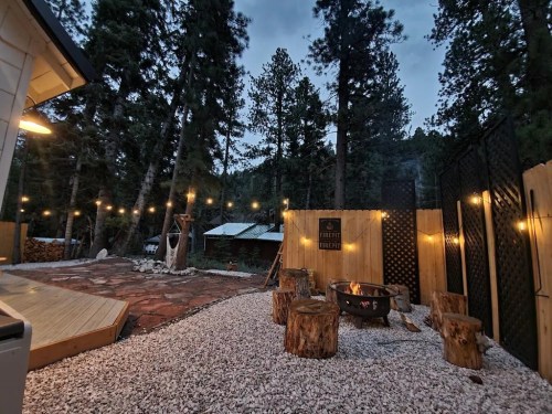 Cozy outdoor space with a fire pit, wooden stools, string lights, and surrounded by tall trees at dusk.