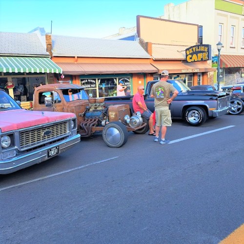 Classic cars lined up on a street, with a man inspecting a vintage vehicle near a café.