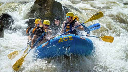 A group of people in a blue raft navigating through whitewater rapids, wearing helmets and paddling.