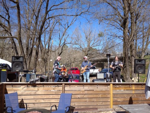 A band performs outdoors on a sunny day, surrounded by trees and equipment, with a wooden deck in the foreground.
