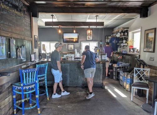 Two people converse at a rustic bar with colorful chairs, shelves of drinks, and a bright, inviting atmosphere.