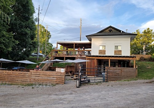 A rustic building with a wooden deck, outdoor seating, and people enjoying the space on a sunny day.