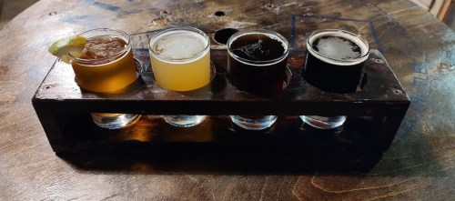 A wooden flight holder with four small glasses of different beers on a rustic table.