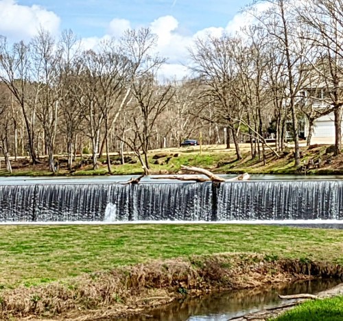 A serene landscape featuring a small waterfall, trees, and a calm river under a blue sky with scattered clouds.