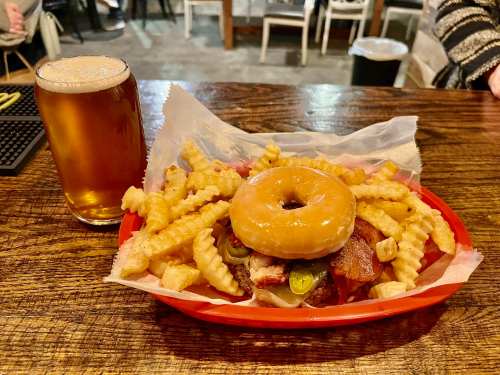 A basket of fries and a sandwich topped with a donut, alongside a glass of beer on a wooden table.