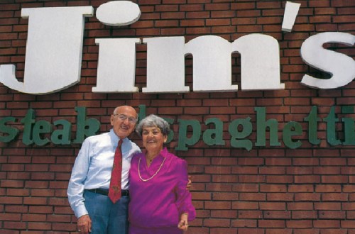An elderly couple smiles together in front of a restaurant sign that reads "Jim's Steak & Spaghetti."