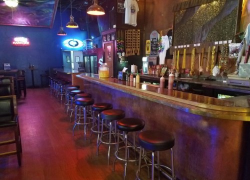 A dimly lit bar with a long counter, stools, and colorful lighting, featuring various drink dispensers and decor.