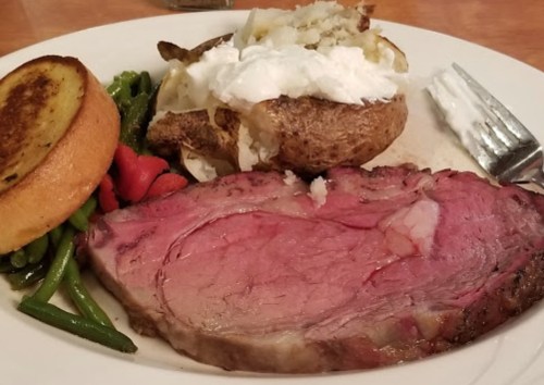 A plate featuring a slice of prime rib, baked potato with sour cream, green beans, and cornbread.