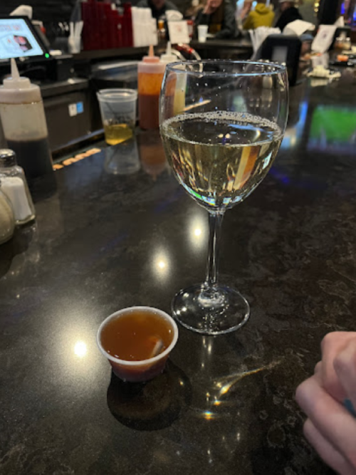 A glass of white wine next to a small cup of brown liquid on a dark bar countertop.