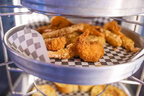 A close-up of a metal tray filled with various fried seafood, including shrimp and fish, on a checkered paper liner.