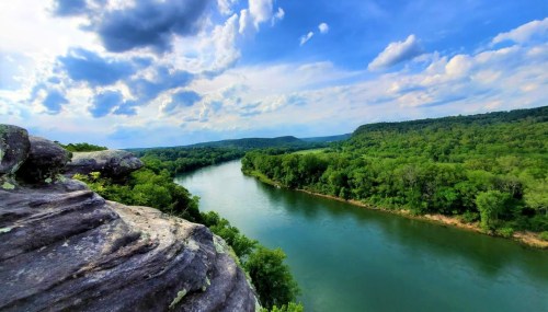 A scenic view of a winding river surrounded by lush green trees and a blue sky with fluffy clouds.