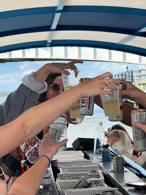 A group of people raising drinks in a toast on a boat, with a scenic waterfront in the background.