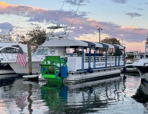 A boat named "Harbor Bar" docked in a marina, reflecting in the water with a colorful sky at sunset.