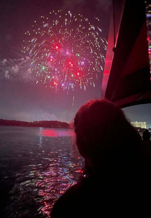 A person watches colorful fireworks explode over a dark lake at night, with reflections shimmering on the water.