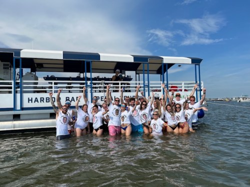 A group of people in matching shirts poses in shallow water near a boat, all smiling and raising their arms in celebration.