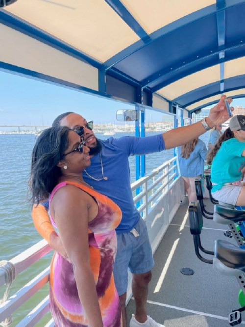 A couple takes a selfie on a boat, enjoying a sunny day by the water with others in the background.