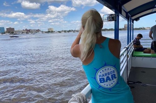 A person in a blue tank top looks out over a river from a boat, with city buildings in the background.