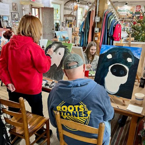 Two people painting at easels in a cozy art studio, with colorful artwork and merchandise in the background.