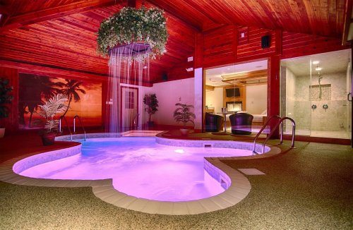 Indoor pool with a heart shape, purple lighting, and a waterfall feature, surrounded by tropical decor and wooden beams.