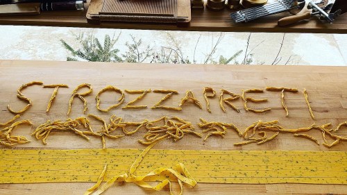Freshly made strozzapreti pasta arranged on a wooden surface, with decorative strands and a sunny outdoor view.