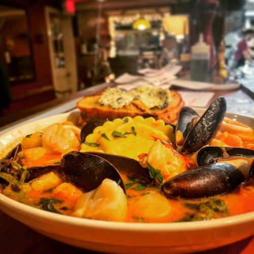 A bowl of seafood pasta with mussels, shrimp, and ravioli, garnished with herbs, served with toasted bread.