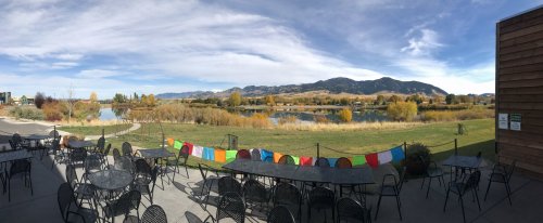 Panoramic view of a serene landscape with a pond, mountains, and colorful flags, framed by outdoor seating.