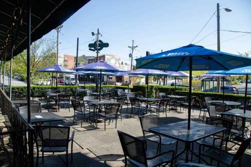 Outdoor dining area with empty tables and blue umbrellas, surrounded by greenery and a street view in the background.