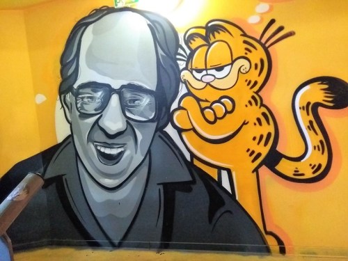 A mural featuring a smiling man in grayscale alongside a cartoon cat, Garfield, on a bright orange wall.