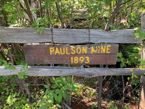 Sign reading "Paulson Mine 1893" attached to a wooden fence, surrounded by greenery.