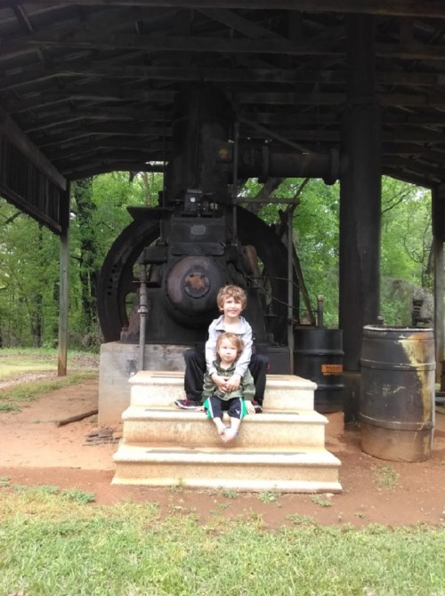Two children sit on stone steps in front of a large vintage machine under a wooden shelter, surrounded by greenery.